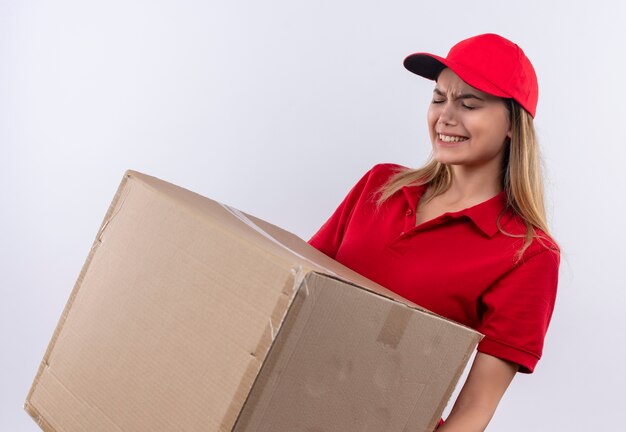 Tense jeune livreuse portant l'uniforme rouge et une casquette tenant une boîte lourde isolé sur blanc
