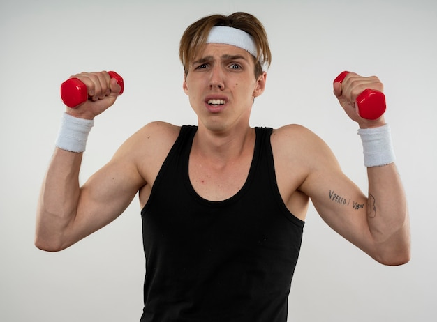 Tense jeune mec sportif portant bandeau et bracelet exercice avec des haltères isolé sur mur blanc