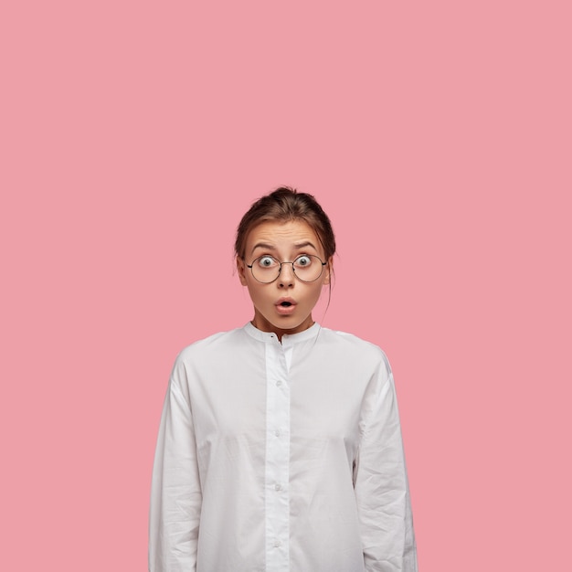 Terrifié jeune femme avec des lunettes posant contre le mur rose