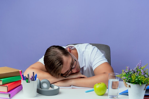 Photo gratuite avec la tête abaissée jeune bel étudiant masculin assis au bureau avec des outils scolaires portant des lunettes