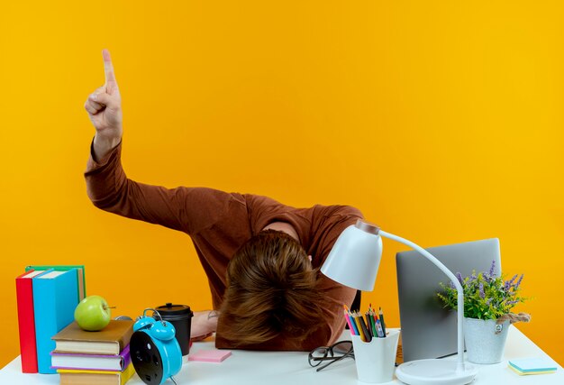 Avec la tête abaissée jeune étudiant garçon assis au bureau avec des outils scolaires points à isolé sur mur jaune avec espace de copie