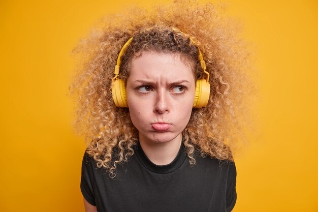 Tête d'une adolescente aux cheveux bouclés mécontente a la mauvaise humeur qui boude l'expression du visage porte des écouteurs stéréo sans fil écoute de la musique vêtue d'un t-shirt noir décontracté isolé sur un mur jaune