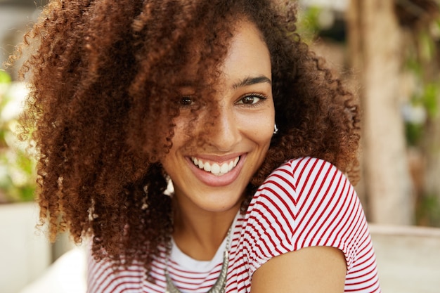 Tête d'adorable belle femme aux cheveux bouclés et au sourire agréable, porte un t-shirt rayé, a des dents blanches même, étant de bonne humeur après un rendez-vous avec son petit ami. Les gens, l'appartenance ethnique et la positivité