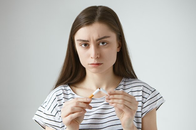 Tête d'autodéterminée sérieuse jeune femme européenne aux cheveux noirs dans des vêtements décontractés tenant deux moitiés de cigarette cassée dans ses mains, arrêter de fumer et briser la mauvaise habitude destructrice