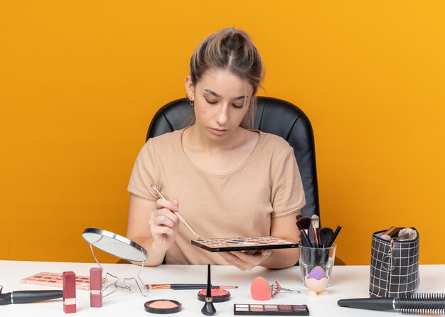 Avec La Tête Baissée, Une Belle Jeune Fille Est Assise à Table Avec Des Outils De Maquillage Appliquant Un Fard à Paupières Avec Un Pinceau De Maquillage Isolé Sur Fond Orange