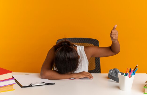 Avec la tête baissée jeune écolière assis au bureau avec des outils scolaires son pouce vers le haut isolé sur un mur orange