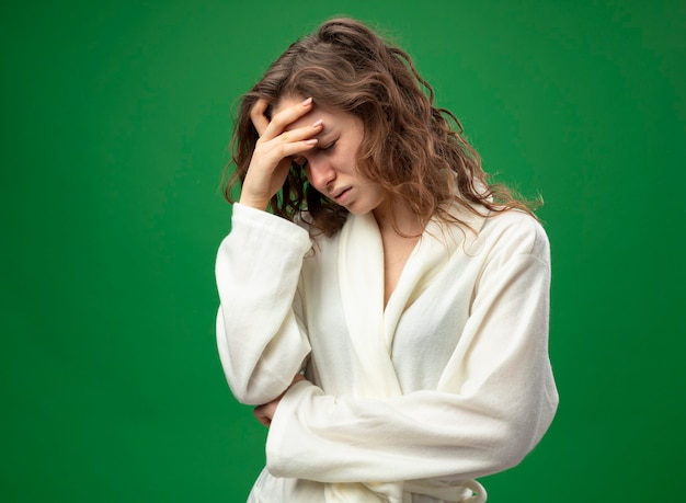 Photo gratuite avec la tête baissée jeune fille malade insatisfaite portant une robe blanche mettant la main sur le front isolé sur vert