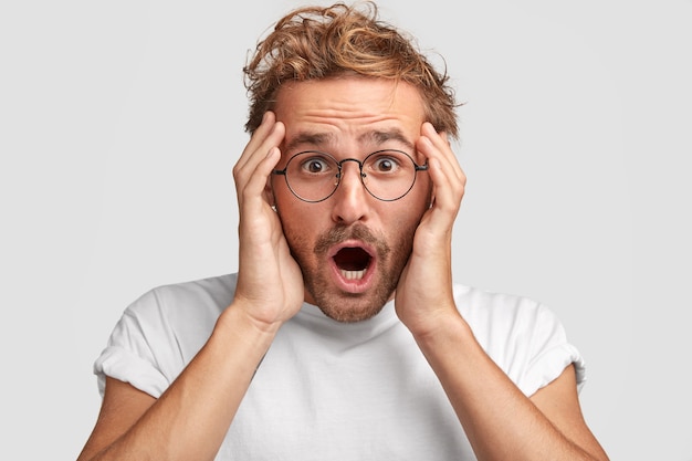 Tête de beau jeune homme mal rasé regarde avec une expression formidable, garde les mains sur la tête, voit comment quelqu'un a écrasé sa voiture, habillé avec désinvolture