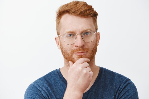 Tête de beau mec rousse créatif et intelligent avec des poils dans des lunettes et un t-shirt bleu, se frottant la barbe sur le menton et regardant avec un sourire narquois, ayant un excellent plan ou une idée sur un mur gris