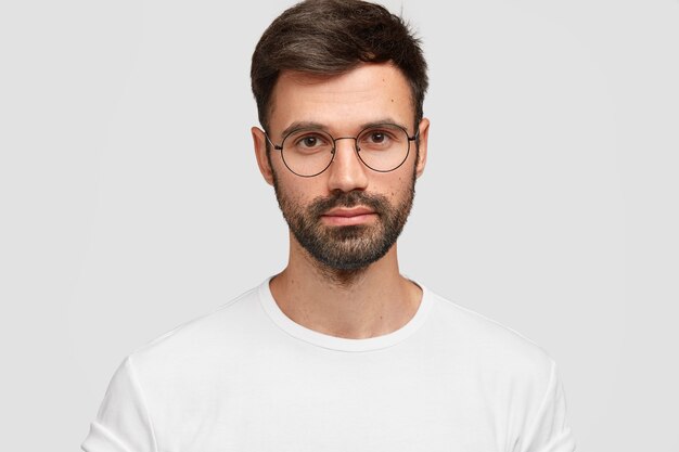 Tête d'un beau pigiste masculin avec un look attrayant, a une barbe et une moustache foncées, regarde directement avec un regard sérieux, porte des vêtements décontractés blancs. Monochrome. Expressions faciales.