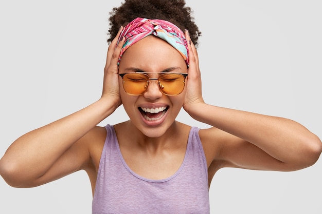 Tête De Belle Femme Afro-américaine Déprimée Hurle Avec Irritation, Garde Les Mains Sur Les Oreilles, Ne Veut Pas Entendre De Mauvaises Nouvelles, Ne Peut Pas Croire En Quelque Chose De Terrible, Isolé Sur Un Mur Blanc