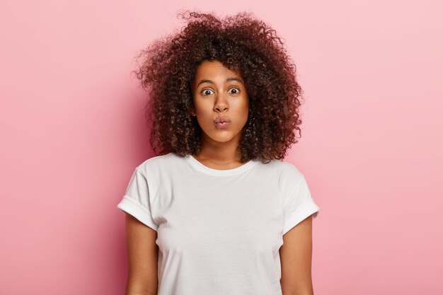 Tête de belle femme à la peau foncée, garde les lèvres pliées, a une coupe de cheveux afro, fait la grimace, a une beauté naturelle, pas de maquillage, porte un t-shirt décontracté blanc, isolé sur un mur rose