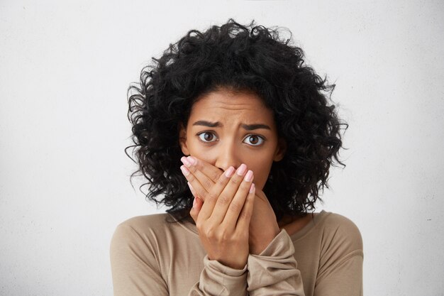 Tête de belle jeune femme européenne à la peau sombre effrayée avec une coiffure frisée fermant la bouche pour ne pas crier, se sentant effrayée et terrifiée, ses yeux et son air pleins de peur et de terreur