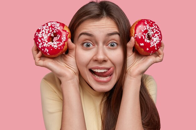 Tête de belle jeune femme lèche les lèvres, regarde avec une expression joyeuse, porte deux délicieux beignets, a les cheveux noirs, apprécie une collation