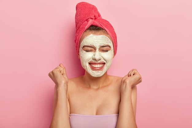 Tête de belle jeune femme lève les poings fermés, applique un masque hydratant sur le visage, a un teint attrayant bien soigné, utilise un produit de beauté naturel pour rafraîchir, enveloppé dans une serviette.