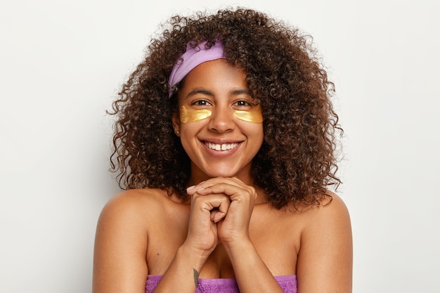 Tête de femme à l'air agréable avec un sourire à pleines dents, cheveux afro touffus, se tient à moitié nue, garde les mains jointes sous le menton, applique des taches jaunes sous les yeux pour la récupération de la peau