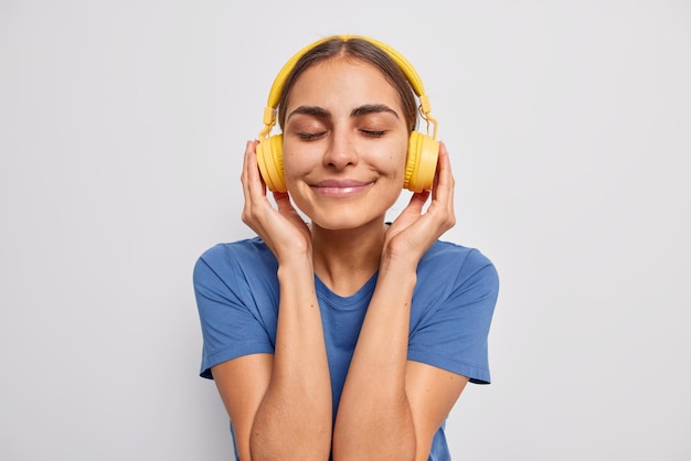 Tête d'une femme à l'apparence agréable ferme les yeux de satisfaction apprécie la musique préférée dans des écouteurs jaunes porte un t-shirt bleu décontracté passe du temps libre sur des poses de passe-temps préférées contre un mur blanc