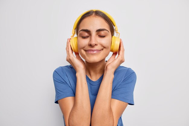 Tête d'une femme à l'apparence agréable ferme les yeux de satisfaction apprécie la musique préférée dans des écouteurs jaunes porte un t-shirt bleu décontracté passe du temps libre sur des poses de passe-temps préférées contre un mur blanc