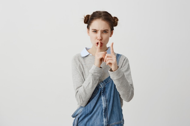 Tête de femme caucasienne grave tenant l'index sur les lèvres. Femme au foyer stricte portant une combinaison de jeans demandant d'être muette parce que ses enfants dorment. Concept de silence