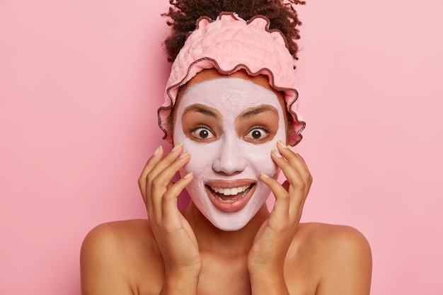 Photo gratuite tête de femme heureuse à la peau sombre applique un masque d'argile, réduit la visibilité des pores, bénéficie d'un résultat efficace du produit de beauté pénétrant profondément dans la peau, se tient à l'intérieur sur un mur rose montre les épaules nues