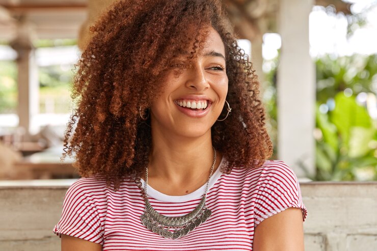 Photo gratuit tête de femme noire drôle aux cheveux bouclés, rit de bonne blague, a un sourire à pleines dents, montre des dents blanches parfaites, porte un t-shirt décontracté à rayures, pose à la cafétéria de la terrasse