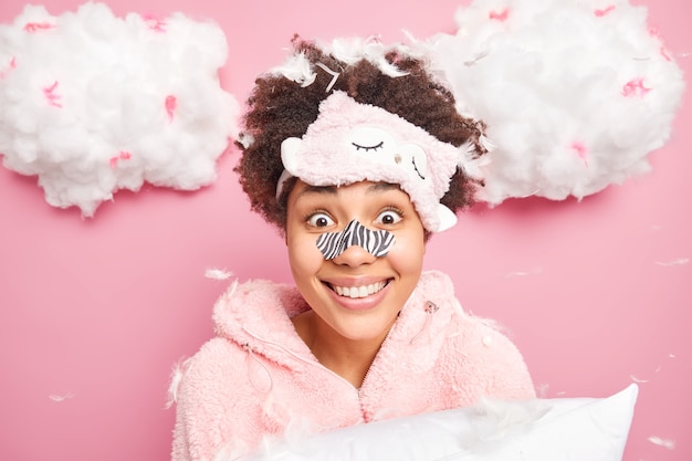 Tête De Femme Positive Aux Cheveux Bouclés Porte Un Costume De Sommeil Avec Patch Nez Bandé Entend Des Nouvelles étonnantes Détient Oreiller Isolé Sur Mur Rose