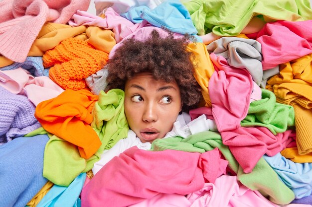 La tête de femme qui dépasse à travers différents vêtements multicolores détourne le regard avec une expression stupéfaite organise le placard purge les vêtements inutiles de la garde-robe. Une femme accro du shopping pose autour d'une tenue