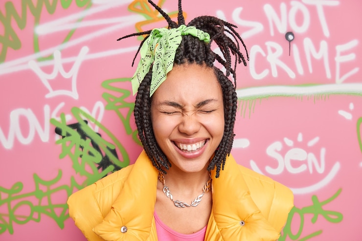 Photo gratuit tête de femme ravie ferme les yeux sourit à pleines dents s'amuse en milieu urbain porte des vêtements élégants pose contre le mur de graffitis de rue exprime des émotions positives