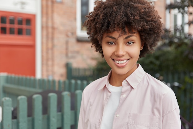 Tête de femme souriante à la peau sombre avec des dents parfaites, se repose en plein air, profite d'une atmosphère calme rurale, regarde directement, habillée avec désinvolture. Gens