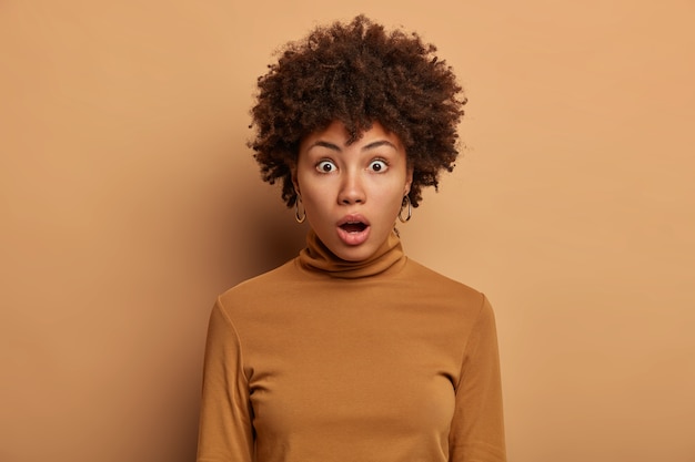 Photo gratuite tête de femme stupéfaite aux cheveux afro, s'interroge sur quelque chose, halète d'excitation, garde la bouche ouverte, vêtue d'un poloneck décontracté, isolée sur un mur beige. certainement pas. peu probable d'être vrai