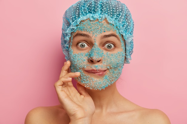 Tête de femme surprise se dresse avec un gommage au sel de mer sur le visage, mord les lèvres et regarde les yeux avec des écouteurs, se sent impressionné par quelque chose, porte un bonnet de bain, affine les pores de la peau, réduit la sécheresse, les taches sombres