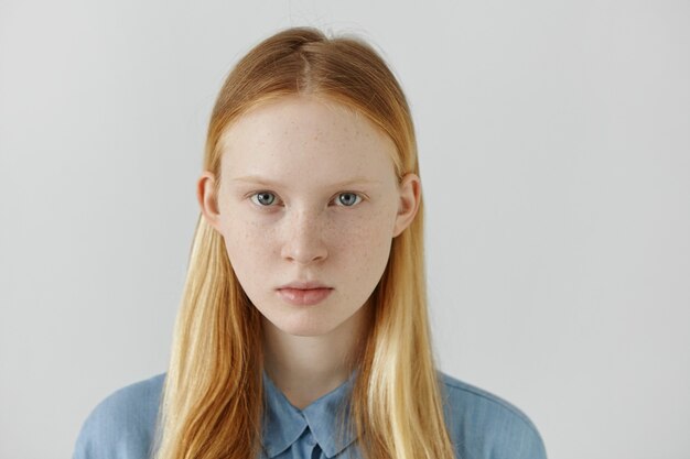 Tête de fille de race blanche avec des taches de rousseur, des cheveux blonds et des yeux clairs vêtus d'une chemise d'école bleue debout contre un mur blanc