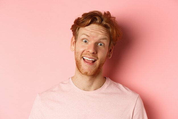 Tête d'un gars rousse drôle montrant la langue, faisant des grimaces idiotes à la caméra, debout joyeux sur fond rose. Espace de copie