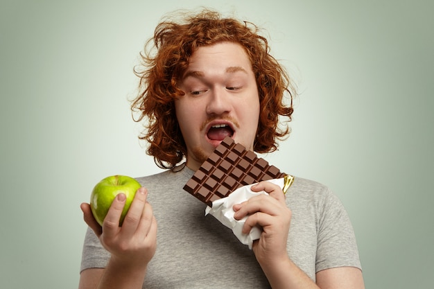 Tête d'un gros homme aux cheveux roux tenant une grosse barre de chocolat dans une main et une pomme verte dans l'autre, choisissant de la malbouffe plutôt que des fruits frais sains, prêts à manger. Les gens et l'obésité