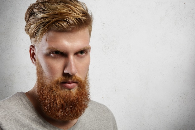 Photo gratuite tête d'homme jeune hipster à la mode avec une barbe épaisse et une coupe de cheveux élégante regardant à distance avec une expression sérieuse et confiante sur son visage.