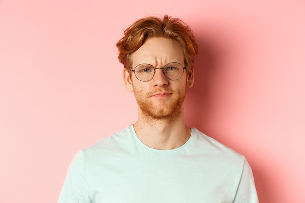 Tête d'homme roux sceptique à lunettes et t-shirt fronçant les sourcils déçu regardant avec désapprobation ...