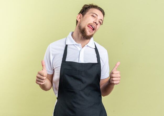 Tête inclinable confiante jeune coiffeur masculin en uniforme montrant les pouces vers le haut isolé sur un mur vert olive