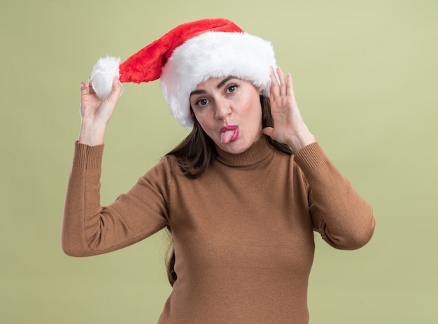 Tête inclinable joyeuse jeune belle fille portant un chapeau de Noël montrant la langue isolée sur mur vert olive