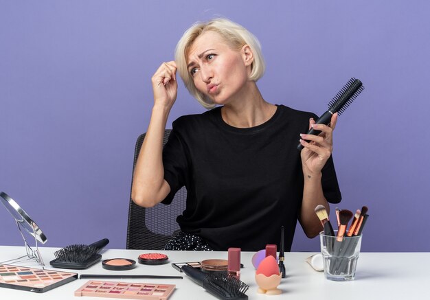 Tête inclinable mécontente jeune belle fille assise à table avec des outils de maquillage tenant un peigne isolé sur fond bleu