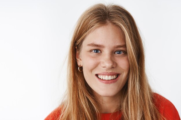 Tête d'une jeune femme européenne charismatique attrayante et insouciante aux yeux bleus profonds, aux cheveux blonds et aux taches de rousseur souriant joyeusement tout en regardant sincèrement et gentil à l'avant debout sur un mur gris