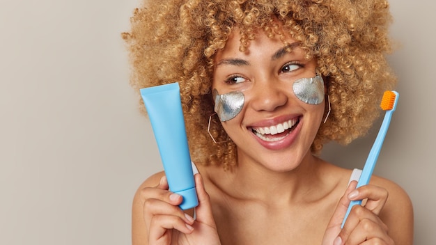 Photo gratuite la tête d'une jeune femme positive aux cheveux bouclés sourit largement montre des dents blanches parfaites tient une brosse à dents et un tube de dentifrice regarde ailleurs joyeusement isolé sur fond gris espace vide pour le texte