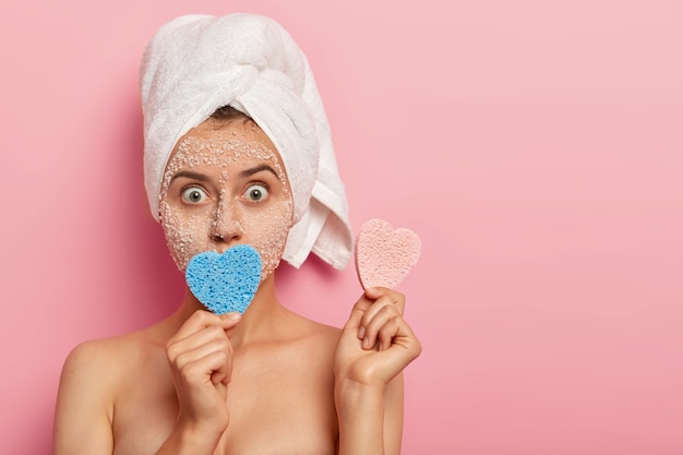 Photo gratuite tête de jeune femme surprise aux yeux bugged, porte une serviette blanche sur la tête mouillée, détient deux éponges en forme de coeur