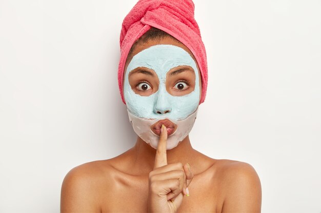 La tête d'une jeune femme surprise fait signe de la main silencieuse, raconte un traitement de beauté, regarde avec des yeux écarquillés, applique un masque à l'argile, un patch en silicone près des lèvres, a une expression mystérieuse, dit être qiet