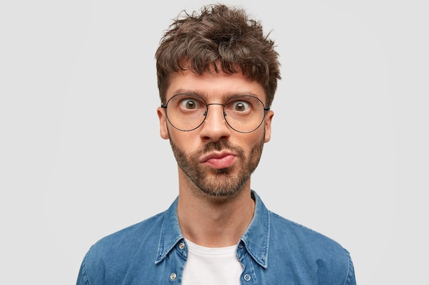 Tête d'un jeune homme barbu perplexe regarde avec émerveillement, courbe les lèvres, a les yeux largement ouverts, les cheveux foncés ondulés, reçoit des nouvelles inattendues, réfléchit à quelque chose