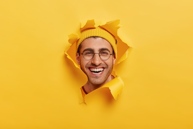Photo gratuite tête de jeune homme non rasé positif sourit largement, porte des lunettes optiques rondes, un couvre-chef jaune