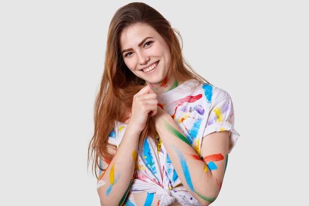 Tête de jeune mannequin femme positive garde les mains ensemble, sourit doucement, porte un t-shirt taché décontracté, aime peindre