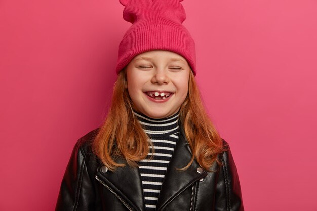 Tête de jolie fille au gingembre a une expression enjouée, ferme les yeux et rit joyeusement, a un sourire positif, se réjouit d'avoir deux dents adultes, va chez le dentiste, isolé sur un mur rose