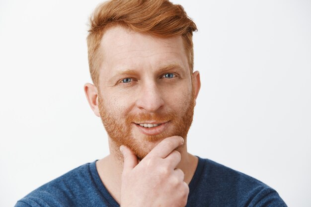 Tête d'un mec rousse attrayant créatif et intrigué avec des poils, se frottant la barbe et souriant, ayant un excellent plan ou une idée regardant curieusement, debout intéressé sur un mur gris