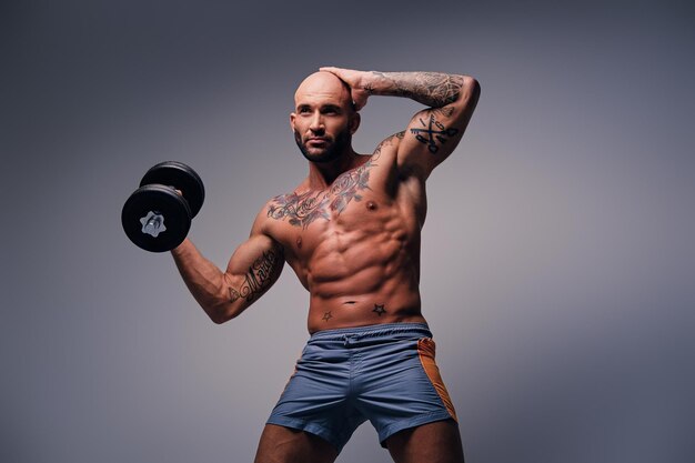 Tête rasée athlétique mâle avec des tatouages sur son torse et ses bras tient une haltère isolée sur fond de vignette grise.