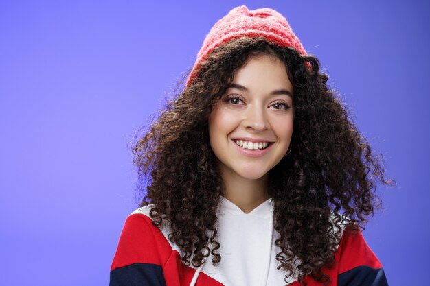 Tête d'une tendre et mignonne femme des années 20 aux cheveux bouclés en bonnet chaud et sweat-shirt cool souriant largement profitant de superbes journées ensoleillées et fraîches à l'extérieur s'amusant à poser sur fond bleu. Espace de copie